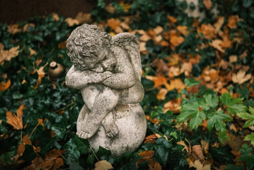 Kamienna figura śpiącego aniołka na tle bluszczu i opadłych jesiennych liści.