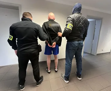 Funkcjonariusze policji eskortują zatrzymanego mężczyznę w kajdankach w korytarzu budynku.