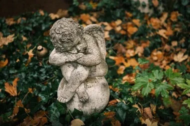Kamienna figura śpiącego aniołka na tle bluszczu i opadłych jesiennych liści.