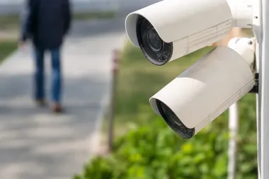 Dwa białe kamery monitoringu zamontowane na zewnątrz, w tle rozmyta postać przechodnia na chodniku.
